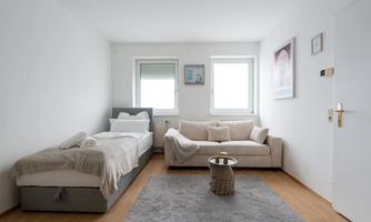 A bright room with a single bed and a sofa. There is a carpet and large windows that let in plenty of daylight.