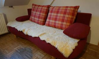 A red sofa with large checked cushions and a soft blanket. The floor is made of wood and the wall is light.