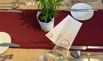 A festively set table with a red table runner and cloth napkins. In the center, there is a small pot with a plant and a menu card.