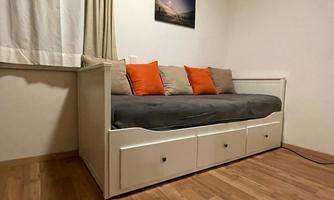 A bright room with a white daybed and orange cushions. A picture hangs on the wall, and the floor is wooden.