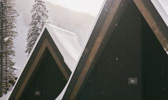 Two snow-covered cabins with pointed roofs in a winter landscape. In the background, there are snow-covered trees and gentle hills.