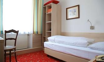A simple hotel room with a single bed, a desk, and a chair. The room has bright windows with colorful curtains and a carpeted floor.