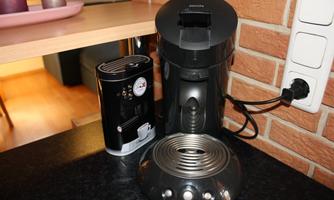 A modern kitchen setup with a coffee machine and a milk frother. The appliances are black and sit on a dark countertop.