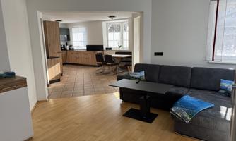A modern living room with a gray sofa and a small table. In the background, there is an open kitchen with wooden furniture and a dining area.