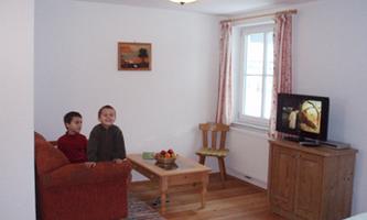 A cozy living room with two children on a couch. There is a television, a table with fruit, and a window with light.