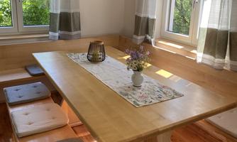 An inviting dining area with a large wooden table and comfortable seat cushions. Large windows let in plenty of daylight and offer a view of the greenery.