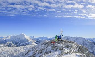 A group of people is standing on a snowy mountain peak and celebrating. The sky is blue with some clouds, and impressive mountain landscapes can be seen in the background.