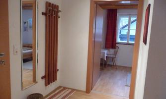 A bright hallway with a mirror and a coat rack. On the right side, there is a table with chairs and a window that lets in natural light.