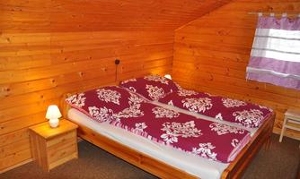 A cozy bedroom with wood paneling and a double bed. The bedding is red with a floral pattern, and there is a nightstand with a small lamp.