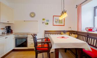 A cozy kitchen with a wooden dining table and chairs. The walls are light, and there are large windows with curtains.