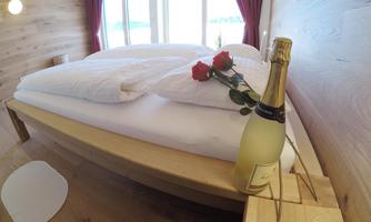 A cozy bedroom with a large bed and two pillows. Next to the bed is a bottle of champagne and a red rose.