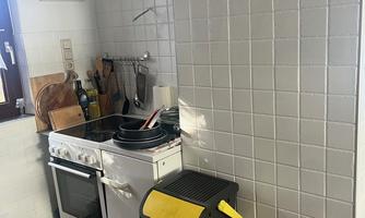 A small kitchen with a stove, a sink, and two trash cans. The walls are covered with white tiles.