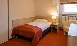 A cozy single room with a bed, a bedside table, and a chair. The walls are light and the floor has a warm carpet.