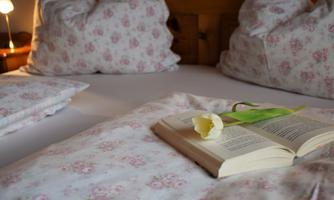 A cozy bed with floral bedding and pillows. On the blanket lies an open book with a flower next to it.