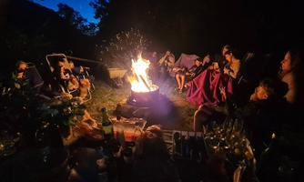 A group of people is sitting around a campfire at night. They are enjoying the atmosphere and sharing drinks and snacks.