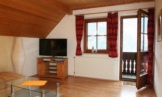 A cozy living room with a wooden beam ceiling and light walls. Windows with red curtains let in plenty of light and provide access to the balcony.