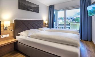 A modern hotel room with two beds and a light wooden floor. Large windows offer a view outside and let in plenty of light.