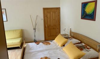 A cozy bedroom with a large bed and yellow pillows. In the background, there is a sofa and a wooden wardrobe.