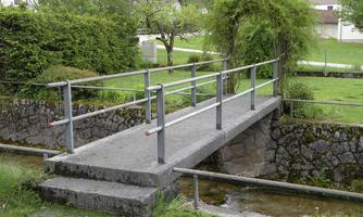 A small concrete bridge with a railing over a narrow creek. Surrounded by green meadows and trees.