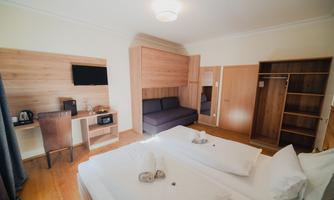A modern hotel room with two single beds and a cozy seating area. The furnishings are made of light wood and include a desk with a television.