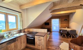 A modern kitchen with wooden shelves and a large window. In the background, there is a cozy living area with a dining table and plenty of natural light.
