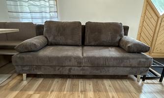 A cozy brown sofa with cushions in a modern living space. The floor is made of wood, and there is a large window area.