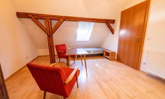 A bright room with a wooden chair and table. The roof is sloped and a large window opening provides plenty of light.