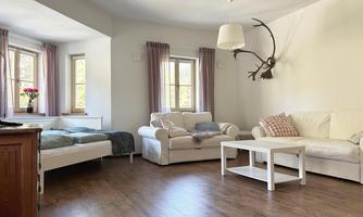A bright living room with a comfortable sofa, a bed, and a white table.  
On the wall hangs a deer antler, and there are large windows with curtains.