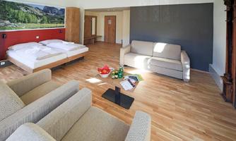 A modern hotel room with two beds, a sofa, and two armchairs. The floor is made of wood and there is a small seating area with magazines and snacks.