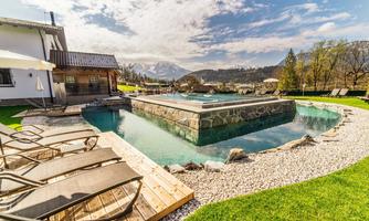 A beautiful outdoor pool with stone cladding, surrounded by lawn and sun loungers. In the background, gentle hills and a clear sky can be seen.