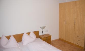 A simple bedroom with a double bed and two large pillows. Next to the bed is a nightstand and a large wooden wardrobe.