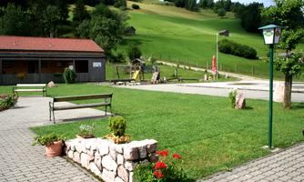 A green meadow with a playground and a bench. In the background, hills and some small buildings can be seen.