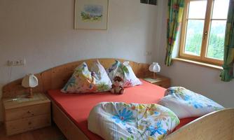 A cozy bedroom with a large bed and colorful bedding. A window lets in plenty of daylight, and a teddy bear sits on the bed.