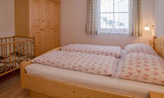 A cozy bedroom with a large bed and a crib for a child. The room has a window with curtains and a wooden wardrobe.
