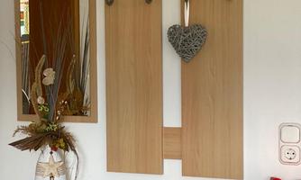 A flat wooden coat rack with several hooks and a heart-shaped decorative element. Next to it stands a vase with decorative branches on a small table.