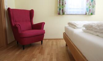 A cozy red armchair is in a bright room. Next to the armchair lies an unmade bed with white bedding.
