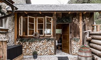 A cozy wooden structure with large windows and a rustic entrance. In front of the building lies a stack of wood and a barrel.