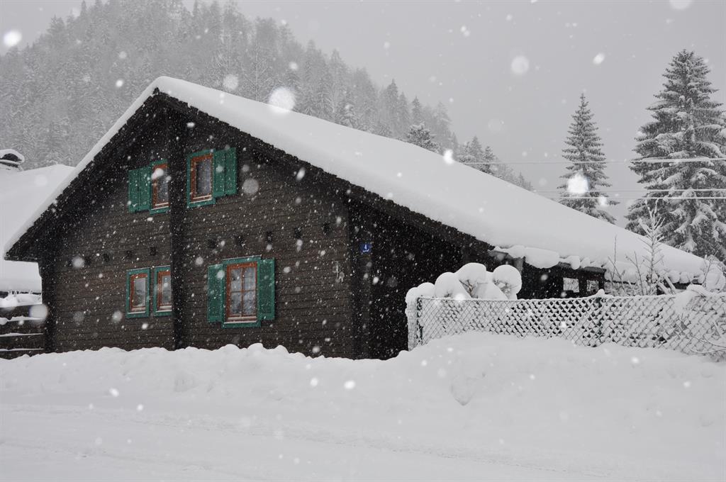 A rustic wooden house in the snow, surrounded by a snowy landscape. The window frames are green and the weather is frosty with falling snow.
