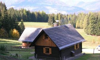 A rustic wooden house in a picturesque landscape with mountains in the background. Surrounded by forests and a green meadow.