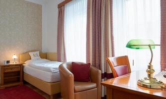 A cozy room with a single bed, a desk, and a comfortable armchair. Large windows provide plenty of daylight.