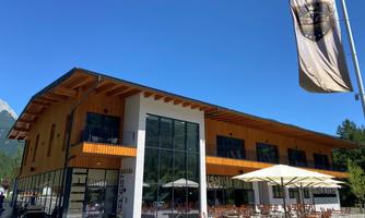 A modern building with large windows and wood siding. Outside, there are tables and umbrellas for guests.