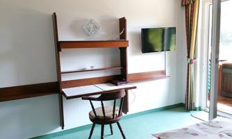 A simply furnished room with a wooden desk and a television on the wall. The room has a balcony with a view outside.