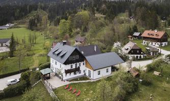 A charming country house surrounded by green nature and mountains. Nearby, additional houses and a quiet road can be seen.