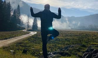 A person is standing on a tree trunk and practicing yoga in nature. In the background, there are mountains and a clear sky visible.