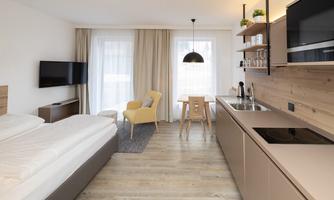 A modern room with two single beds, a television, and a small kitchen. There is a dining table and a cozy armchair next to large windows with curtains.