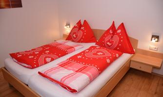 A cozy bedroom with a double bed and red bedspreads. The walls are light and the lighting is soft.