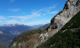 An impressive mountain landscape with steep rocks and green forests. The sky is clear and blue, and in the background, more mountains are visible.