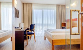 A modern hotel room with two beds and a desk.  
Large windows provide plenty of daylight and a view outside.