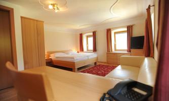 A bright and spacious room with a double bed and a cozy seating area. There are windows with curtains and a television that create an inviting atmosphere.