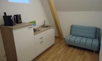 A small kitchenette with a refrigerator and a kettle. In the room, there is a blue sofa and light wooden flooring.